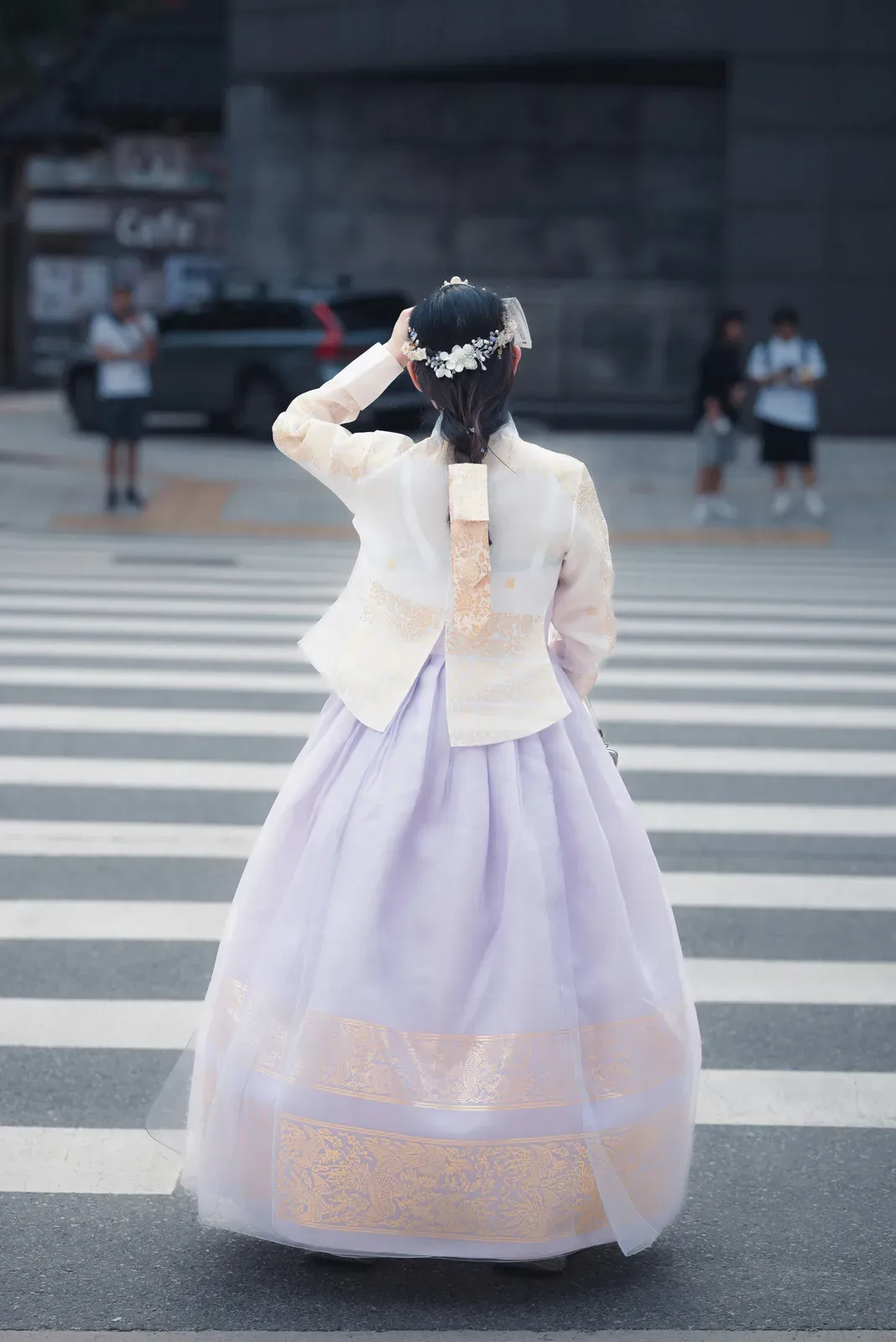 Woman seen from behind crossing a zebra crosswalk in Seoul in a pastel hanbok with a cream jacket, a full lavender skirt and flowers in her hair
